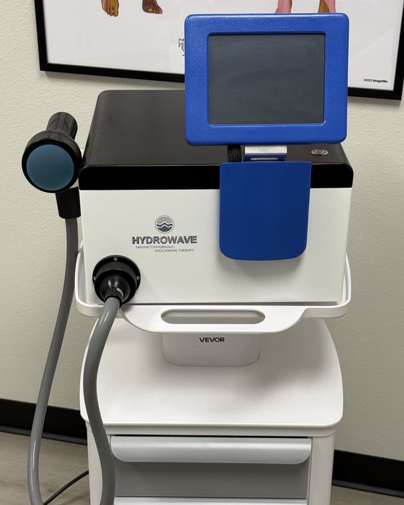 The Hydrowave shockwave unit in the Tailored Touch office: a white console with a blue touchscreen and a handheld applicator on a grey hose, mounted on a rolling cart